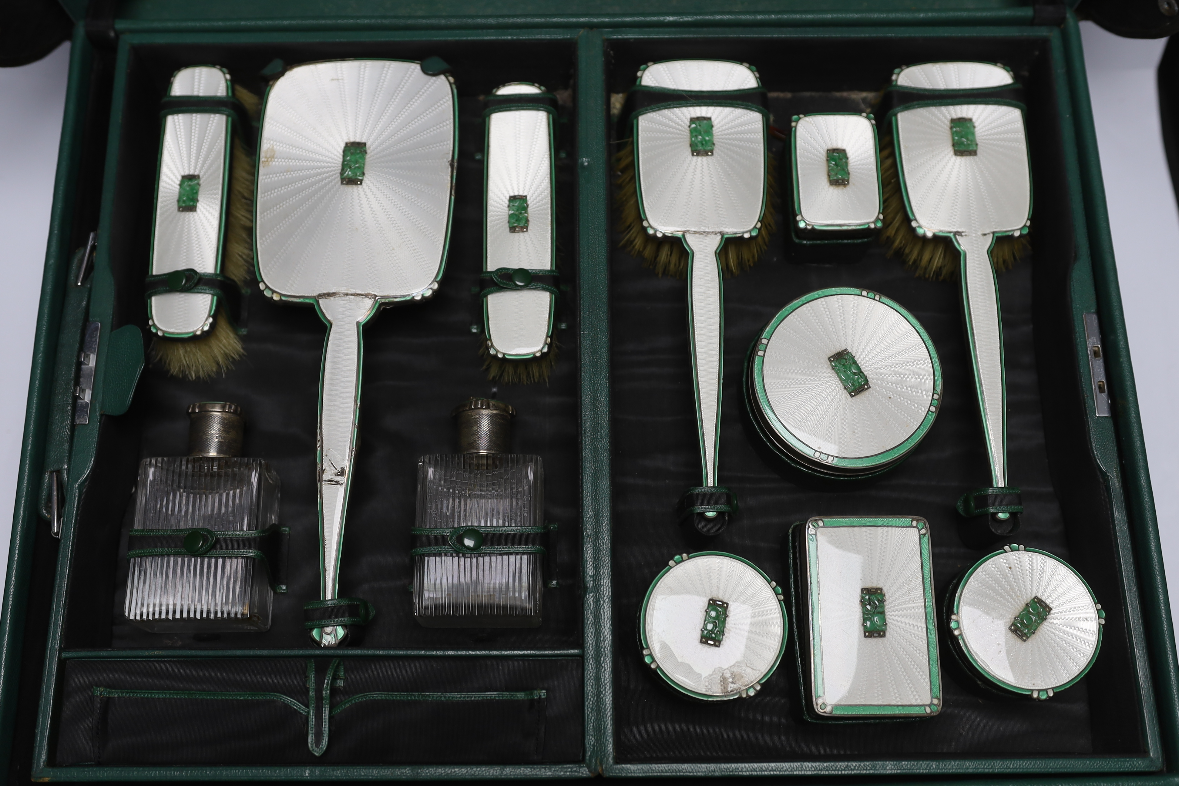 A George V silver, enamel and Chinese jadeite mounted twelve piece dressing table set, Goldsmiths & Silversmiths Co Ltd, Birmingham, 1936, housed in green leather travelling case, case width 53cm (a.f.).